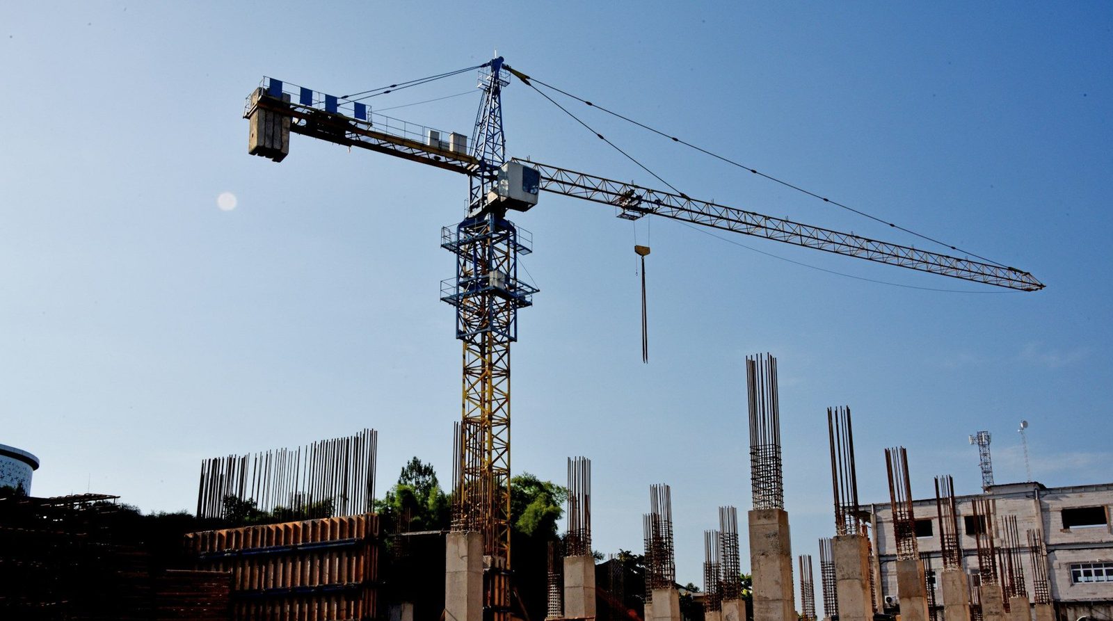 Tower crane rising above new concrete columns and rebar on a construction site under a clear blue sky