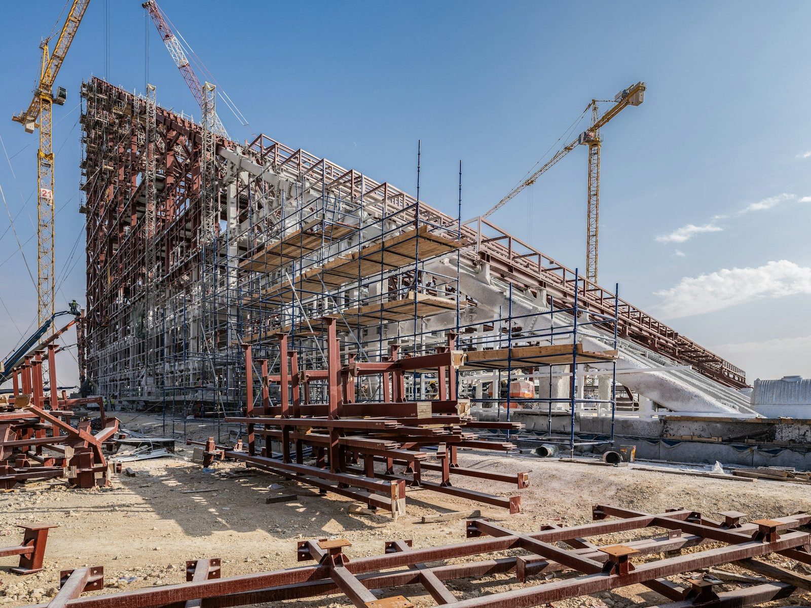 Steel-frame industrial structure under construction rising from bare ground, with tower cranes against a clear blue sky