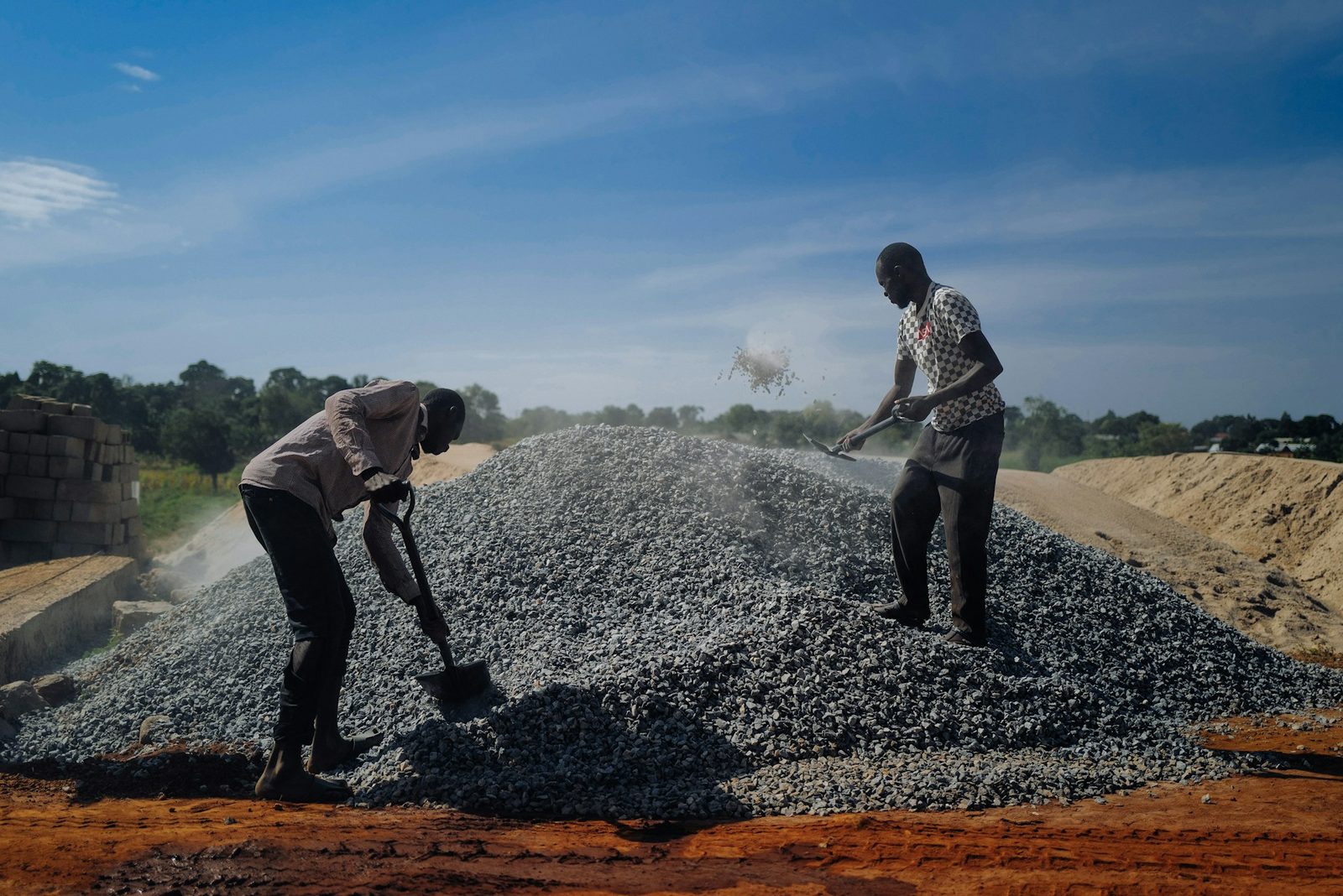 Two African workers shoveling crushed stone under a bright blue sky