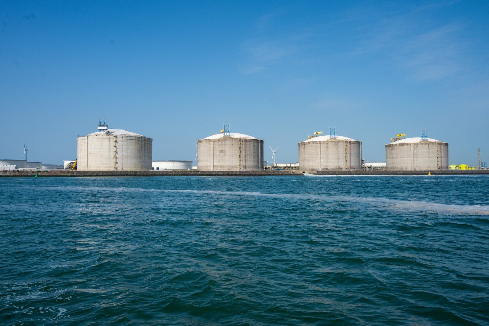 Four large fuel storage tanks on a waterfront terminal under a clear blue sky with wind turbines behind