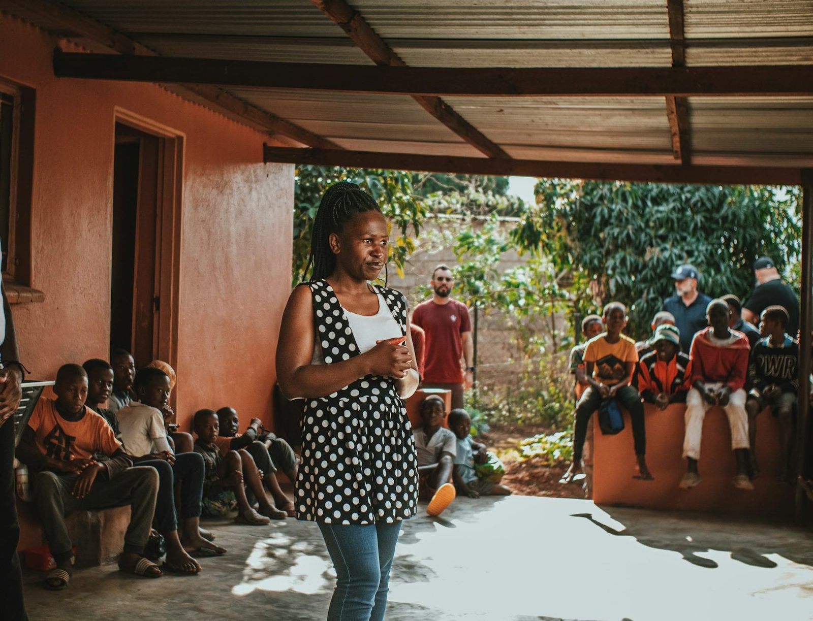 Woman facilitator addresses a seated community meeting under a porch in rural Africa